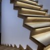 Spiralne stopnie z jasnego drewna, podświetlone od spodu ciepłym światłem, przylegające do białej, zakrzywionej ściany.