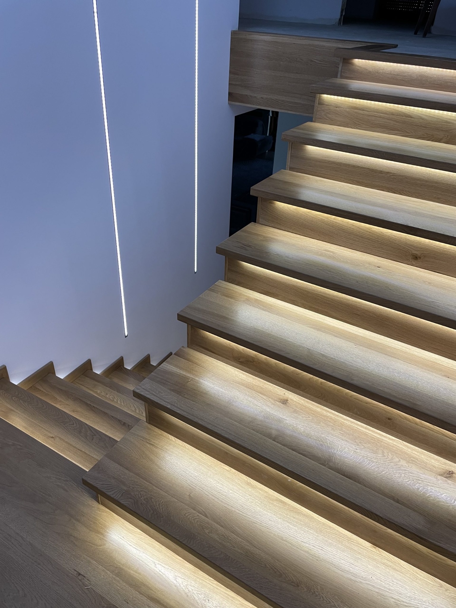 Nowoczesne schody z drewna dębowego z podświetleniem LED, minimalistyczny design, biała ściana z pionowymi lampami.
