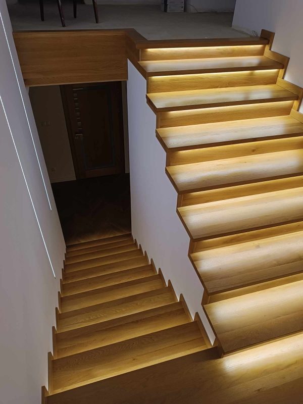 Drewniane schody z podświetleniem LED wzdłuż stopni, widok z góry, kontrastujące z ciemnym wejściem i minimalistycznym oświetleniem ściennym.