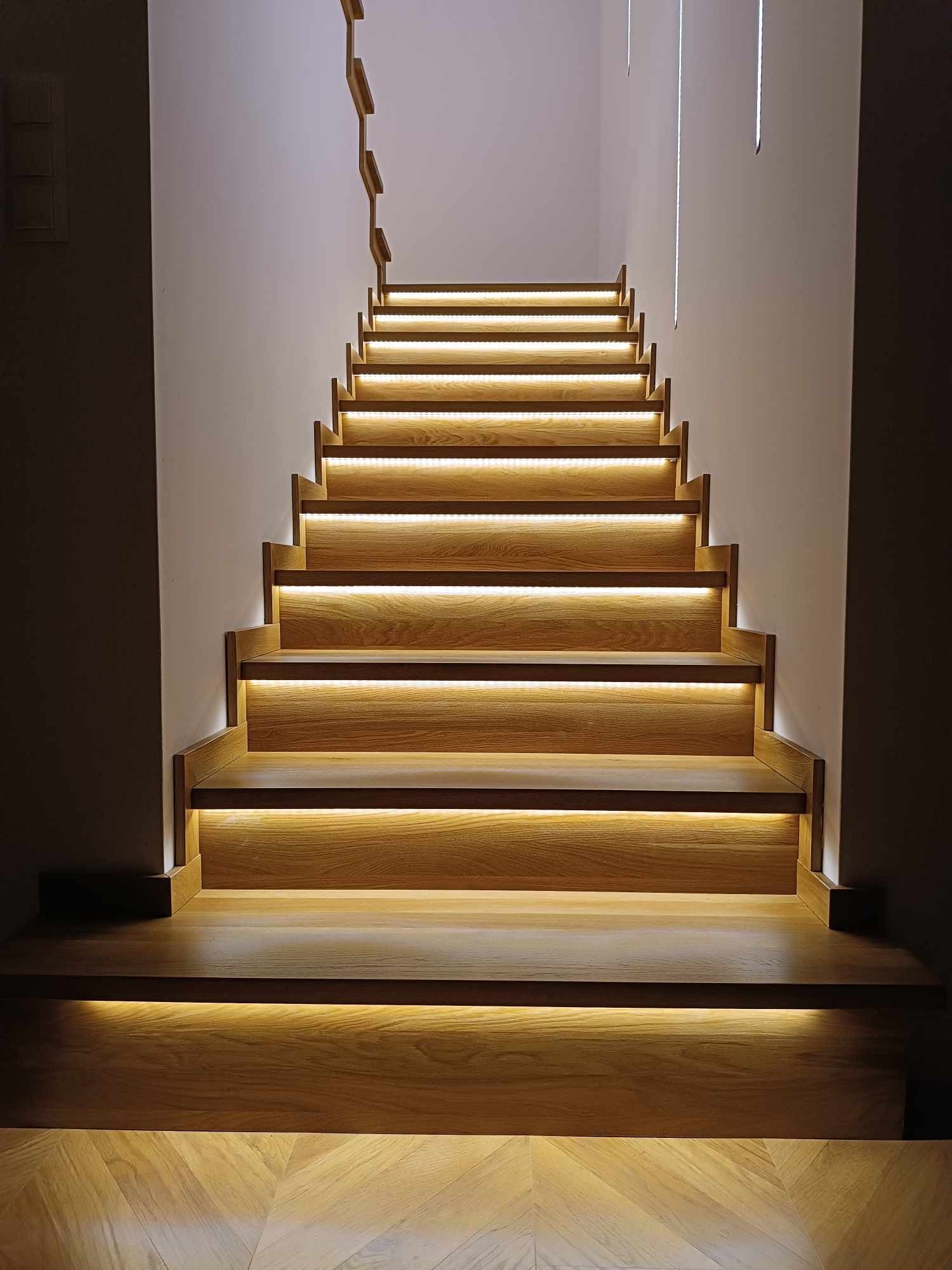 Nowoczesne schody z jasnego drewna z podświetleniem LED na każdym stopniu, widok od dołu, z elementami dekoracyjnymi na ścianie.