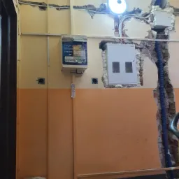 Ściana w trakcie remontu z widocznymi przewodami elektrycznymi, puszkami i skrzynką bezpiecznikową, częściowo zabezpieczonymi pianką montażową, z uszkodzonym tynkiem i fragmentami cegieł.