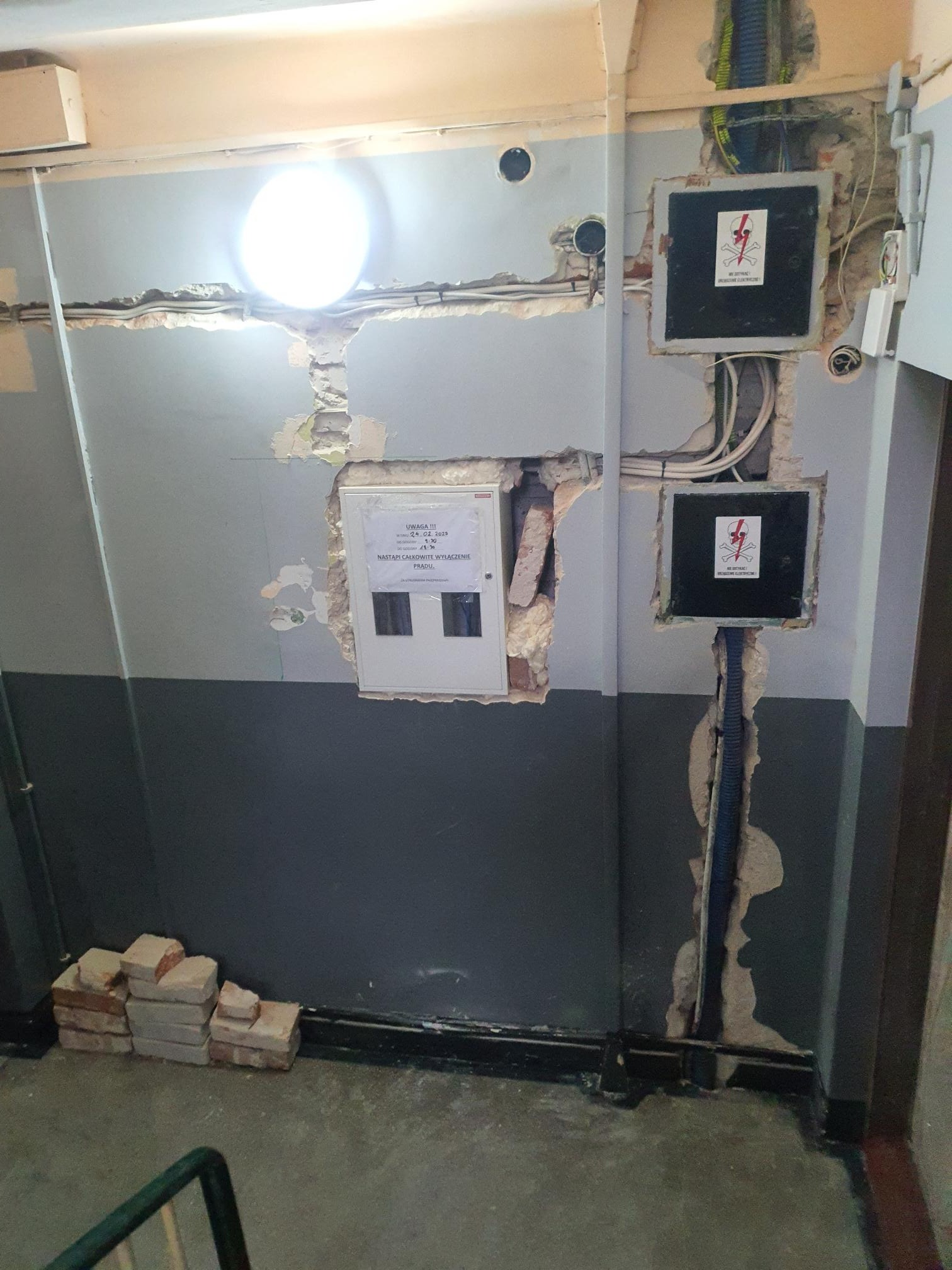 Klatka schodowa w trakcie remontu z odsłoniętymi przewodami elektrycznymi, puszkami i tablicą informacyjną o wyłączeniu prądu; widoczne uszkodzenia ściany i cegły na podłodze.