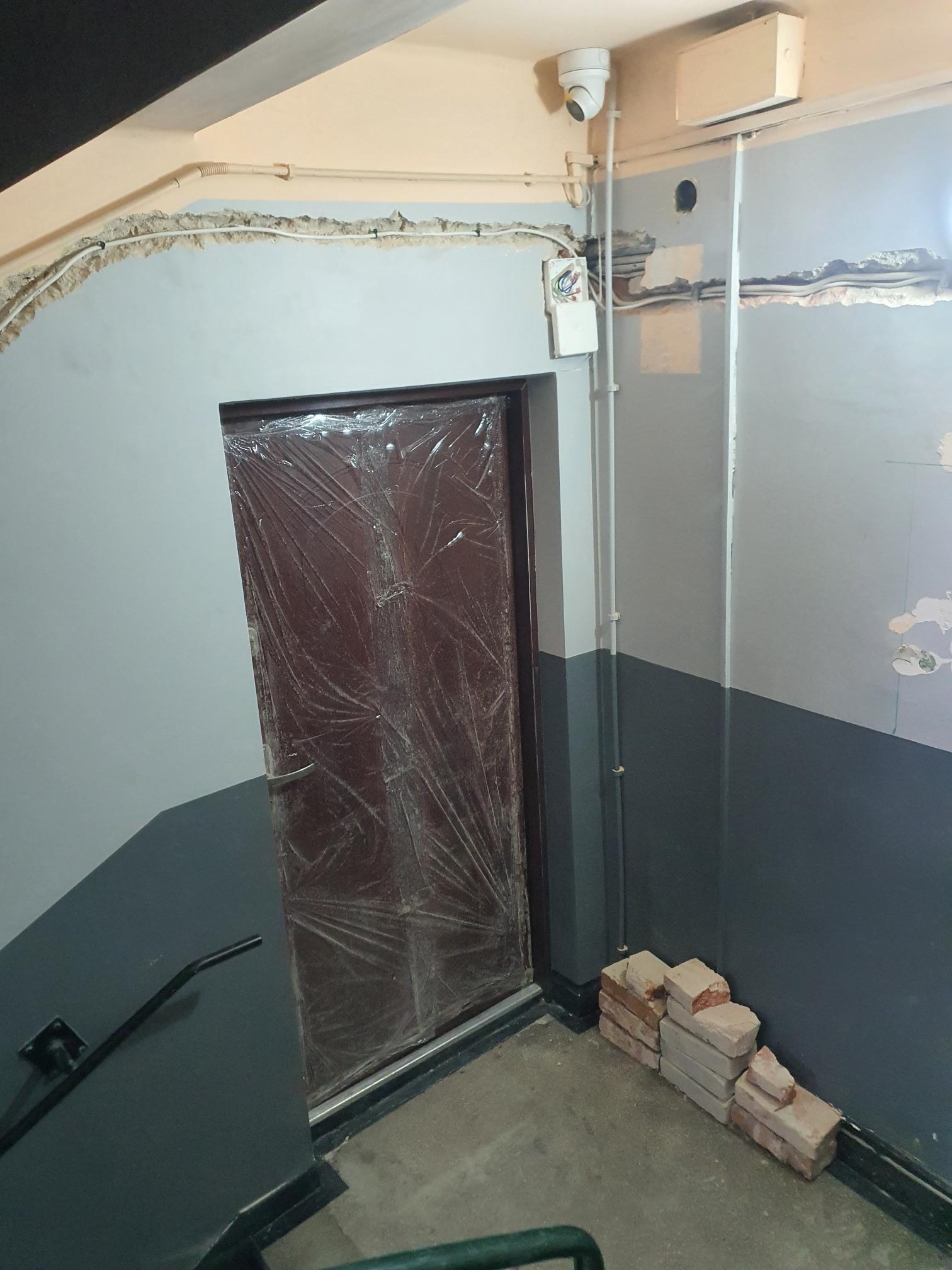 Korytarz w trakcie remontu: widoczne przewody elektryczne pod sufitem, kamera monitoringu, zabezpieczone folią drzwi, ślady po kuciu w ścianie i stos cegieł przy podłodze.