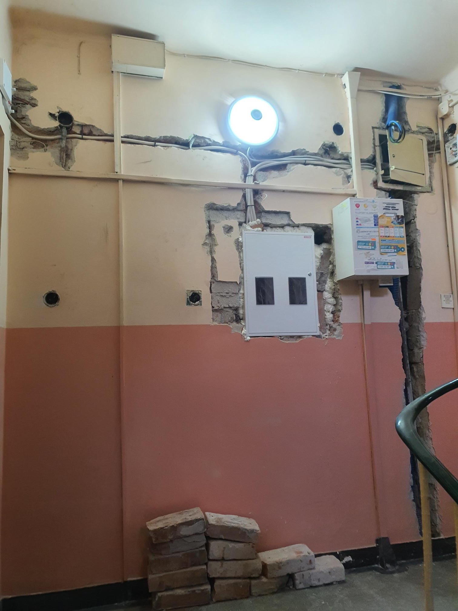 Montaż elektryczny: skrzynka rozdzielcza z dwoma okienkami, licznik energii i przewody w ścianie z widocznymi uszkodzeniami tynku i stertą cegieł na podłodze.
