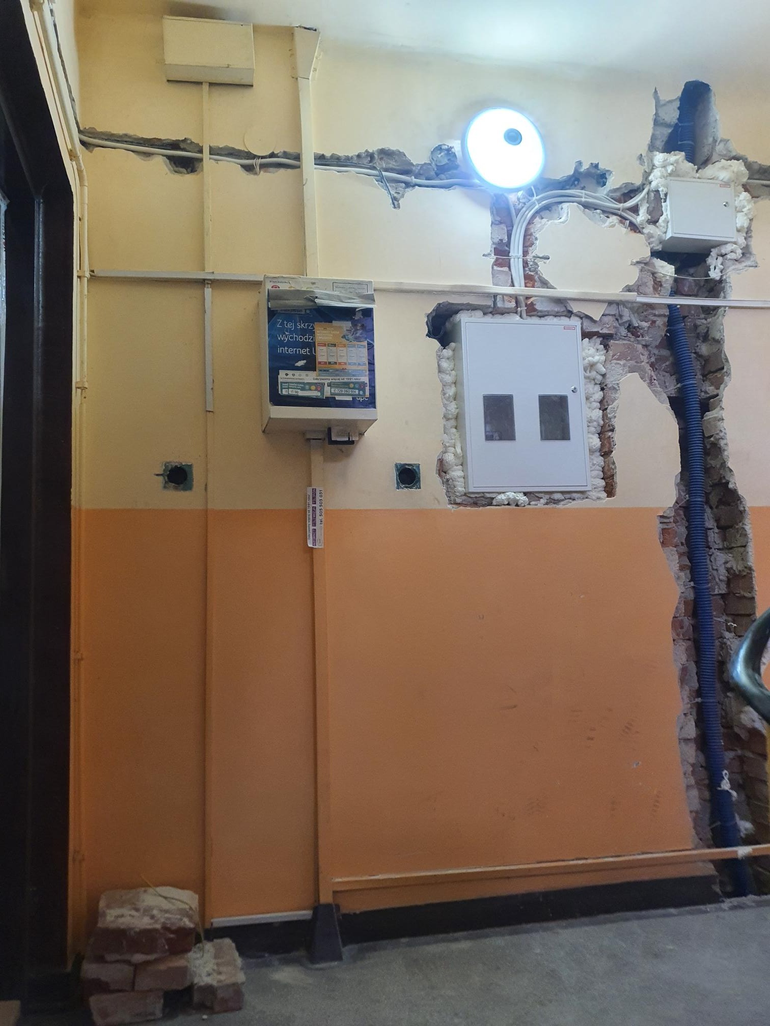 Ściana w trakcie remontu z widocznymi przewodami elektrycznymi, puszkami i skrzynką bezpiecznikową, częściowo zabezpieczonymi pianką montażową, z uszkodzonym tynkiem i fragmentami cegieł.