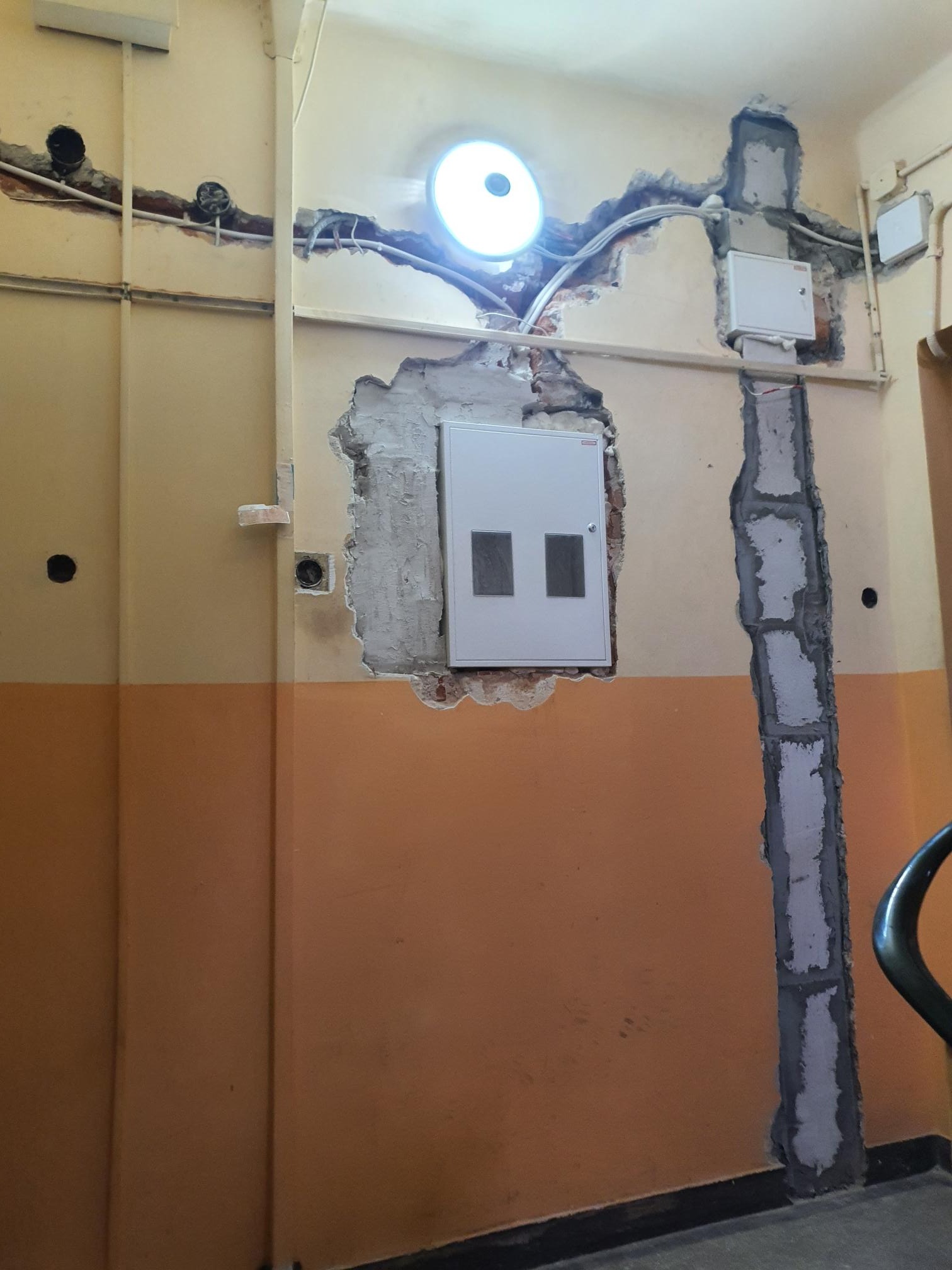 Wnętrze w trakcie remontu z odsłoniętymi przewodami elektrycznymi w ścianach, widoczne puszki instalacyjne i skrzynka bezpiecznikowa, ślady po kuciu w ścianie, lampa sufitowa oświetla miejsce prac.