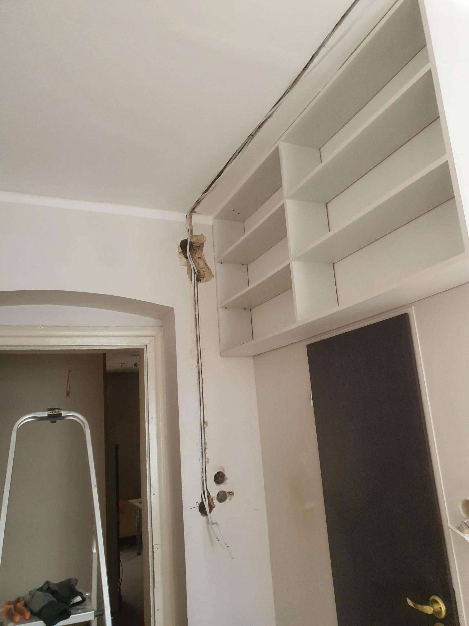Wnętrze w trakcie remontu: widoczne przewody elektryczne wystające ze ściany obok białych półek i drzwi, drabina w tle, przygotowane do prac wykończeniowych.