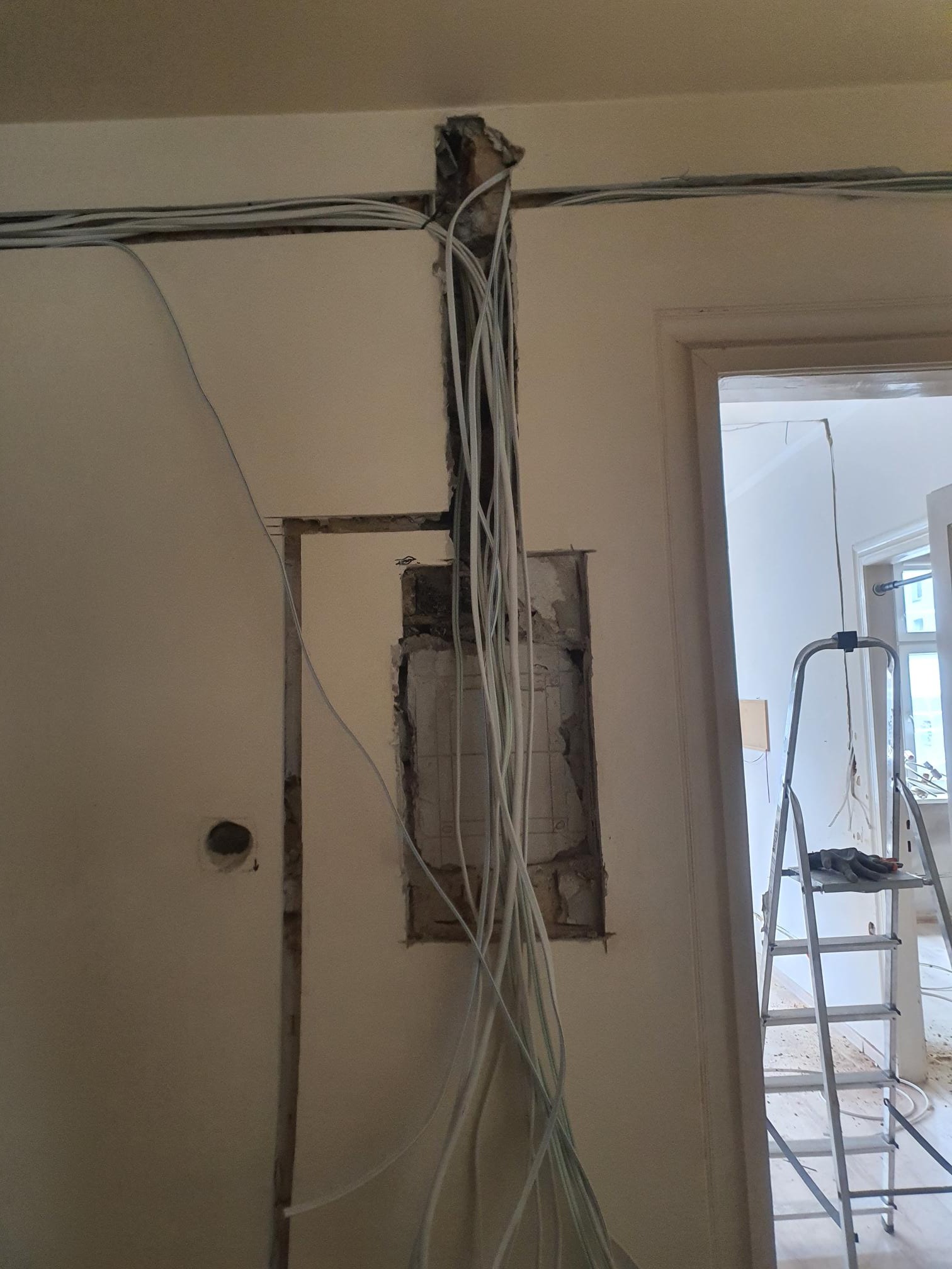 Ściana z wyciętymi bruzdami i otworem na puszkę elektryczną, widoczne kable w peszlach, drabina aluminiowa w tle, wnętrze w trakcie remontu.