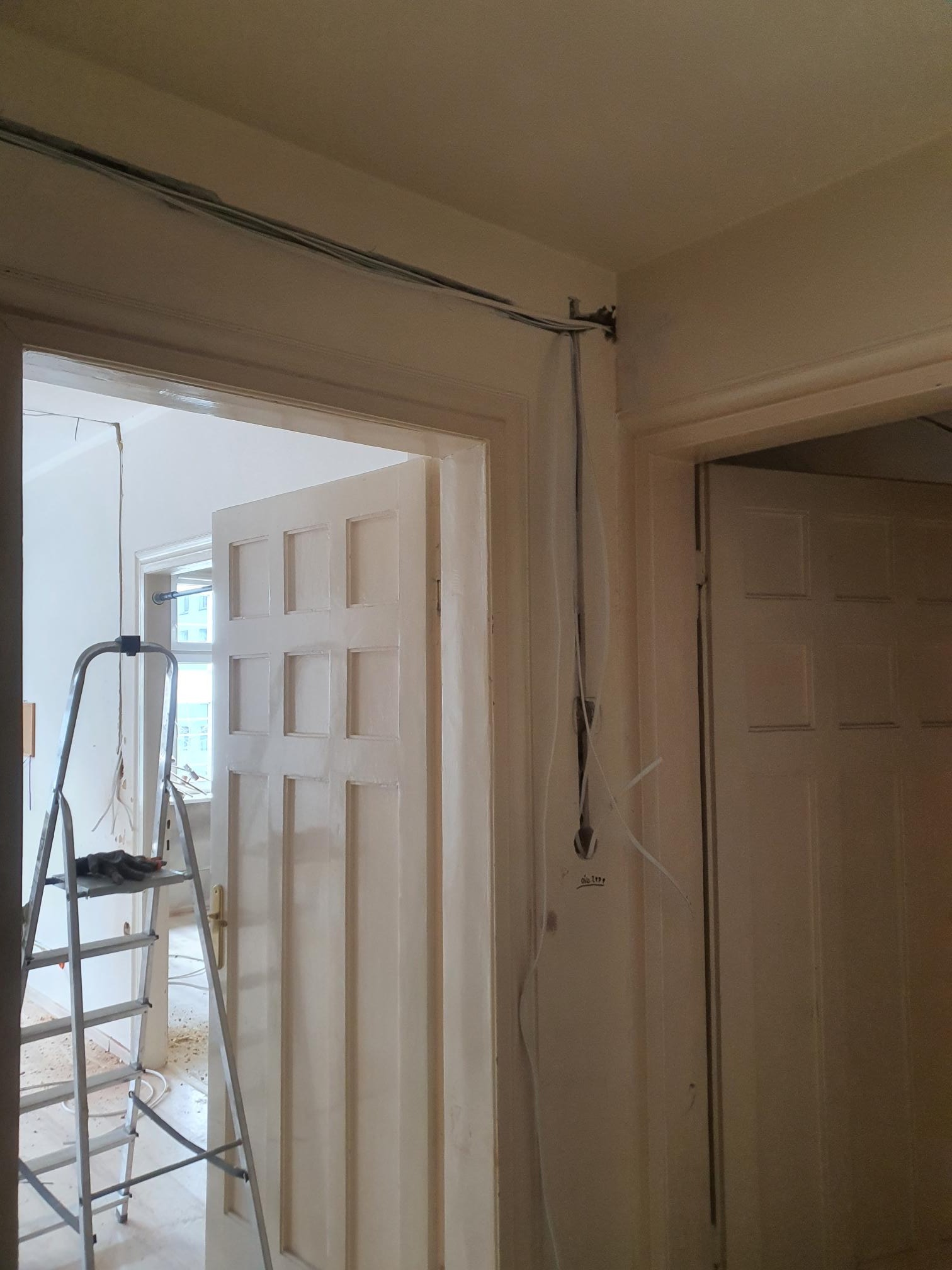 Wnętrze w trakcie remontu: widoczne kable elektryczne prowadzone po ścianie, drabina aluminiowa w tle, otwarte drzwi z widokiem na okno.