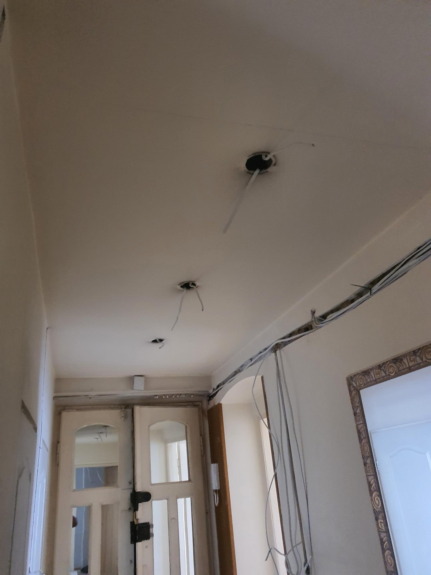 Wnętrze w trakcie remontu: widoczne kable wystające z otworów w suficie, listwa z przewodami przy ścianie, stare drzwi z szybami.
