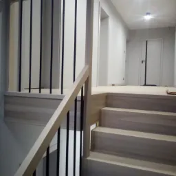 Jasne drewniane schody z czarnymi metalowymi prętami balustrady, widok z dołu, minimalistyczny design, białe ściany i drzwi w tle.