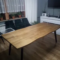 Stół z blatem z jasnego drewna o nieregularnych krawędziach, wsparty na czterech czarnych nogach, na tle okna z białymi firankami i ciemnozielonych krzeseł.