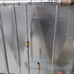 Metalowe rury instalacyjne z zaworami i złączkami na tle blachy trapezowej, widoczne elementy instalacji grzewczej i hydraulicznej.