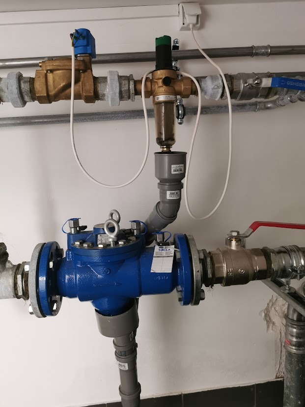 Złożony system rur hydraulicznych z różnymi zaworami i filtrem, widoczne złącza kołnierzowe i rury PCV, instalacja przy białej ścianie.