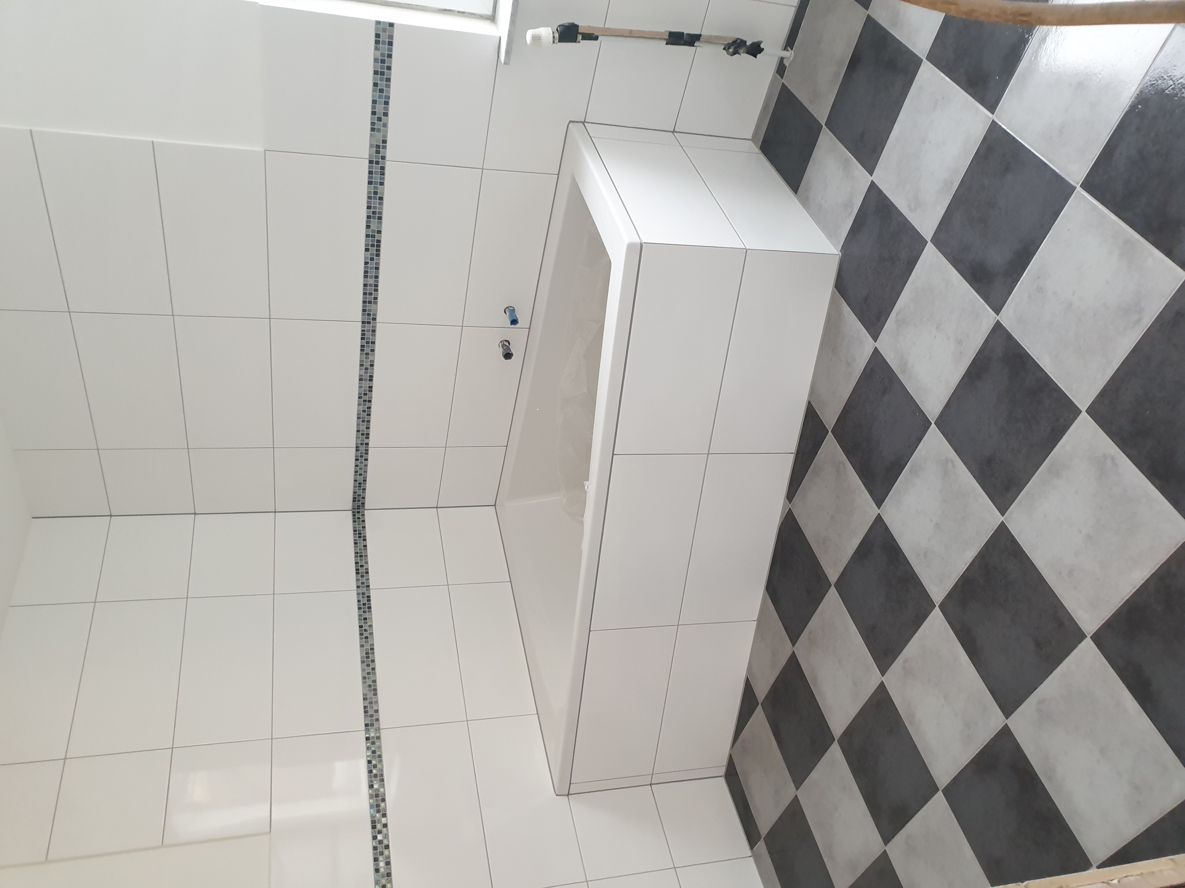 Wykończona łazienka w Korzybiu z białą wanną obudowaną płytkami, białe płytki na ścianach z dekoracyjnym paskiem mozaiki i podłoga wyłożona czarno-białą szachownicą.