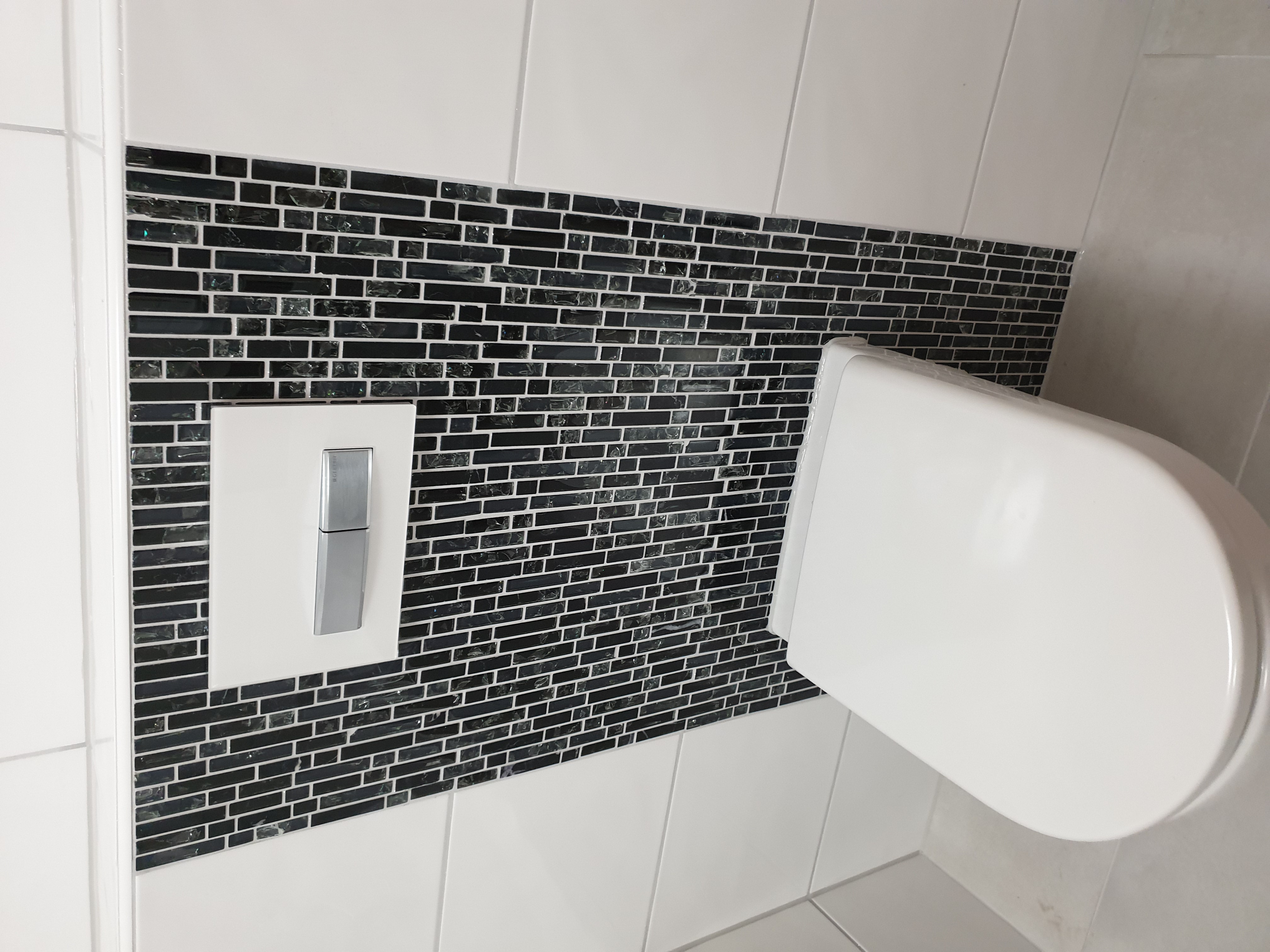 Ściana w łazience wyłożona białymi i czarnymi, błyszczącymi, prostokątnymi płytkami mozaikowymi, z widocznym przyciskiem spłuczki i fragmentem toalety w kolorze białym.