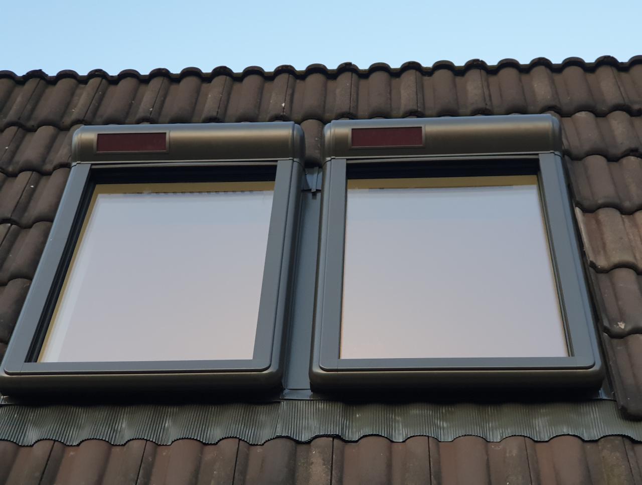 Dwa okna dachowe z zewnętrznymi roletami w kolorze grafitowym, zamontowane na brązowym dachu z dachówki ceramicznej.
