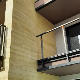 Elewacja budynku wielorodzinnego wykończona panelami imitującymi drewno, z widocznymi balkonami z szklanymi balustradami i elementami montażowymi w folii ochronnej.