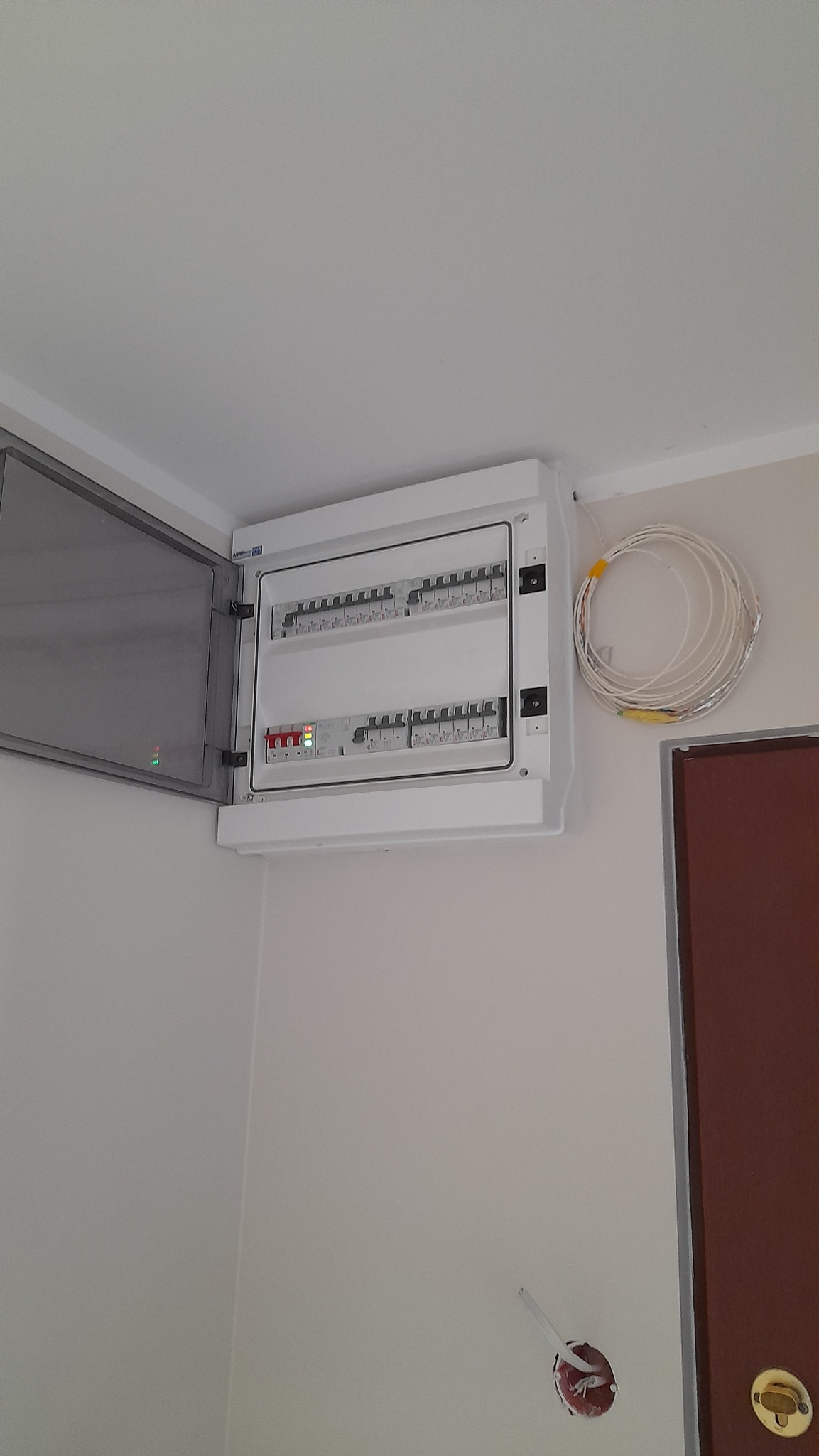 Rozdzielnica elektryczna z otwartymi drzwiczkami zamontowana na ścianie, obok zwój kabli i puszka instalacyjna.