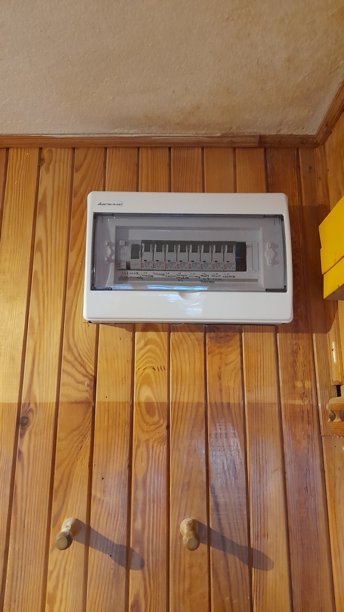 Zamknięta biała skrzynka elektryczna Elektro-Plast zamontowana na ścianie z boazerii, widoczne bezpieczniki wewnątrz.