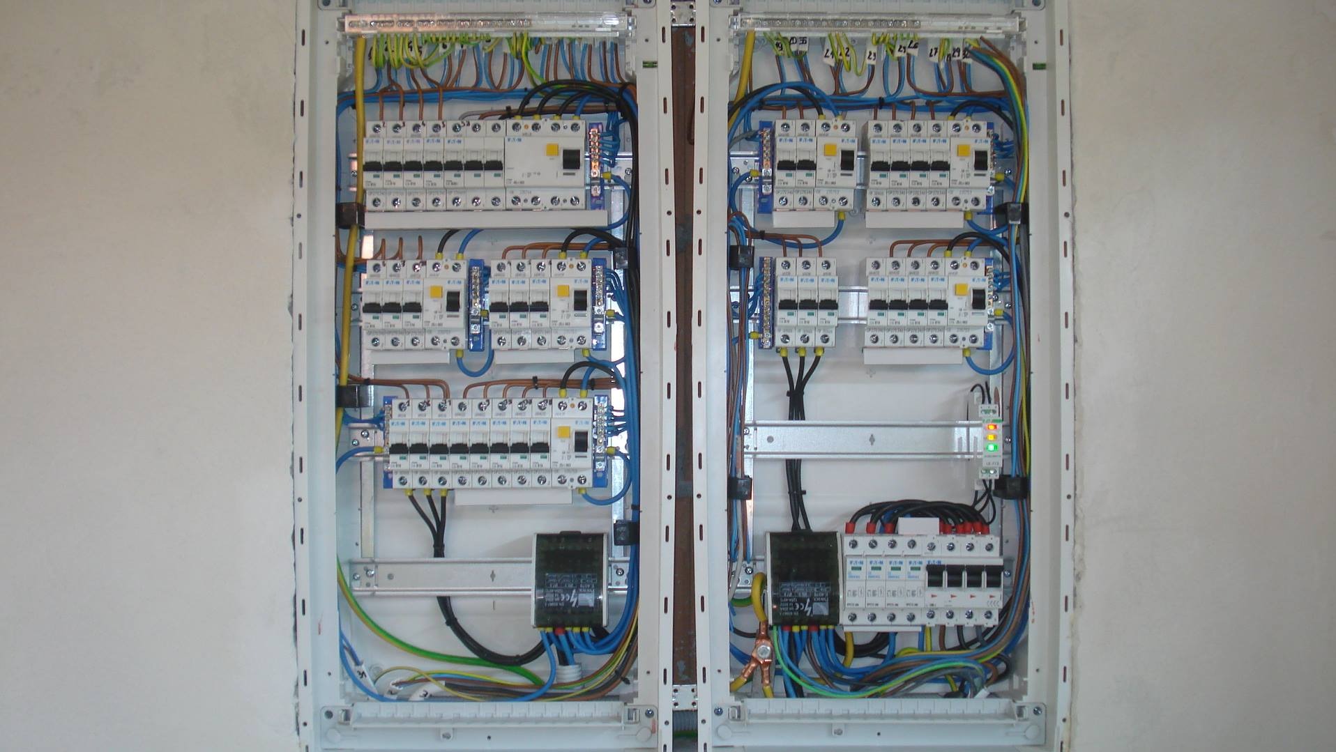 Rozdzielnica elektryczna dwudrzwiowa, z widocznymi bezpiecznikami, przewodami w różnych kolorach i elementami montażowymi, zamontowana na szarej ścianie.