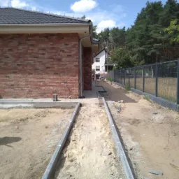 Budowa ścieżki z obrzeżami betonowymi przy murowanym domu z czerwonej cegły, widoczna metalowa siatka ogrodzeniowa i niebieski pojemnik na wodę.