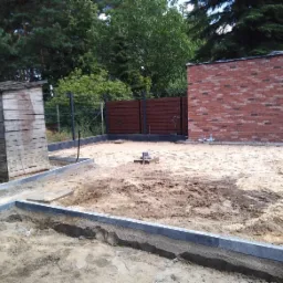 Przygotowanie terenu pod budowę: wytyczone fundamenty z obrzeżami, częściowo wymurowana ściana z cegły klinkierowej, drewniana szopa w tle.
