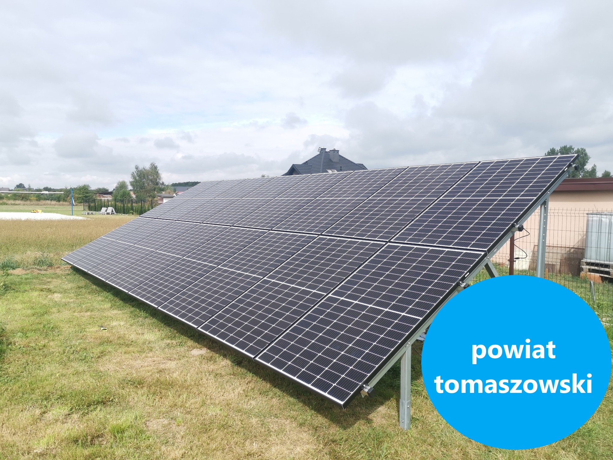 Panel słoneczny na metalowej konstrukcji, zainstalowany na trawiastym terenie, z widocznym domem w tle i napisem 'powiat tomaszowski' w niebieskim okręgu na pierwszym planie.