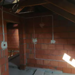 Wnętrze poddasza w trakcie prac, widoczne ceglane ściany z rozprowadzonymi kablami elektrycznymi, puszki elektryczne zamurowane w ścianie, drewniane elementy konstrukcyjne dachu.