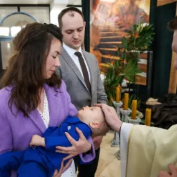 Matka trzyma śpiącego syna w niebieskim ubranku podczas ceremonii religijnej chrztu, ksiądz dotyka głowy dziecka, a ojciec stoi obok w szarym garniturze, widoczne świece i dekoracje w tle.