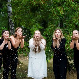 Fotografia Katarzyna Guzicka - Pięć kobiet w plenerze dmucha konfetti z dłoni, centralna postać w białej sukience, pozostałe w czarnych strojach, tło stanowią zielone drzewa.