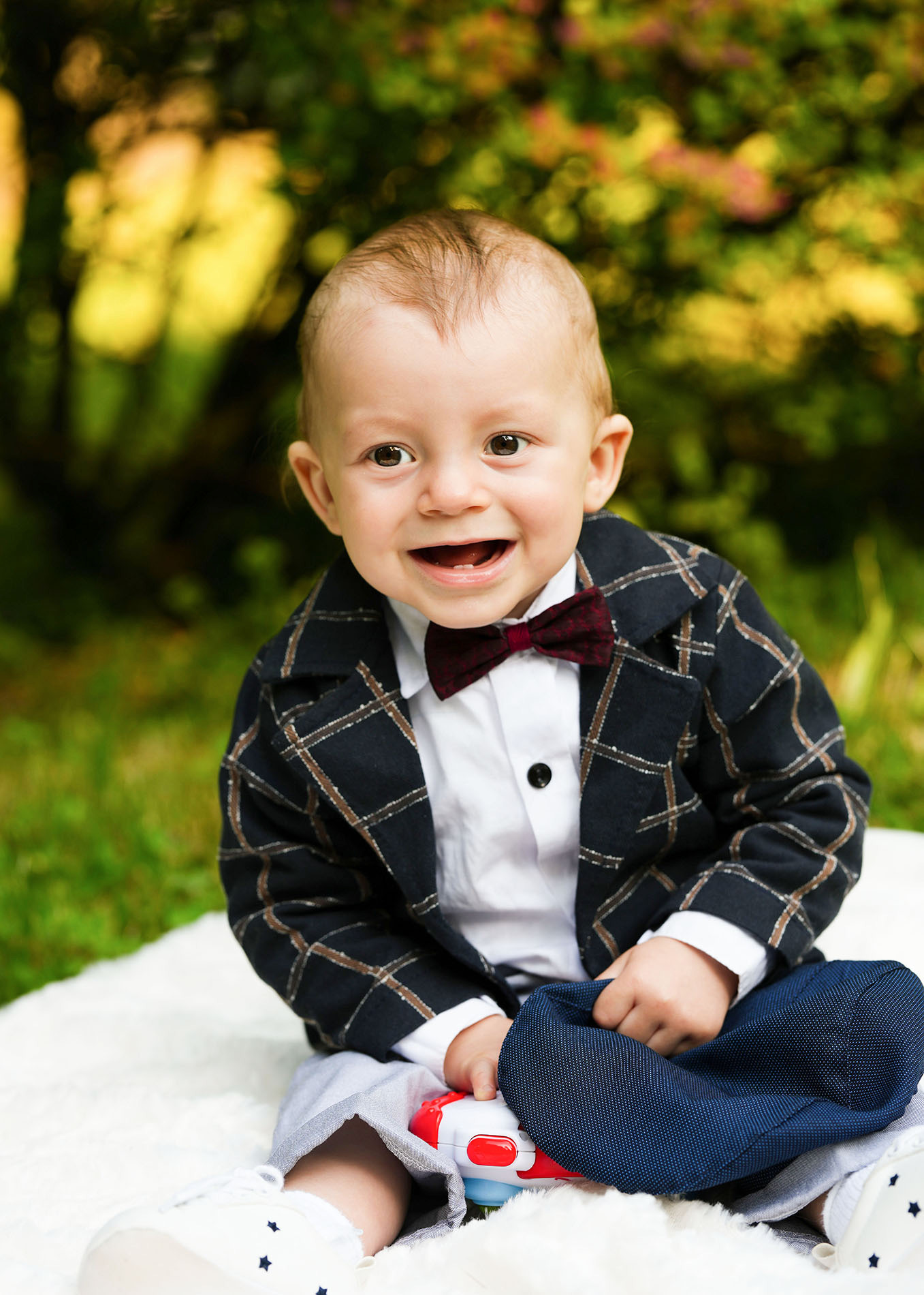 Uśmiechnięty niemowlak w eleganckim ubranku, siedzący na puszystym kocyku na tle zieleni, trzymający w rękach zabawkę.