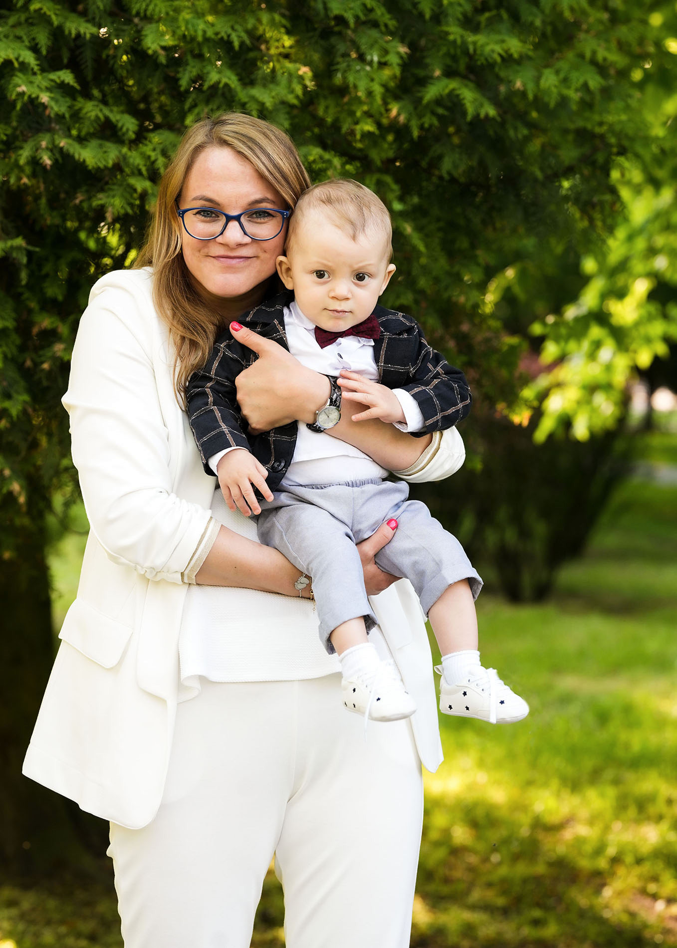 Mama w białym garniturze trzyma elegancko ubranego synka z muszką na tle zieleni.