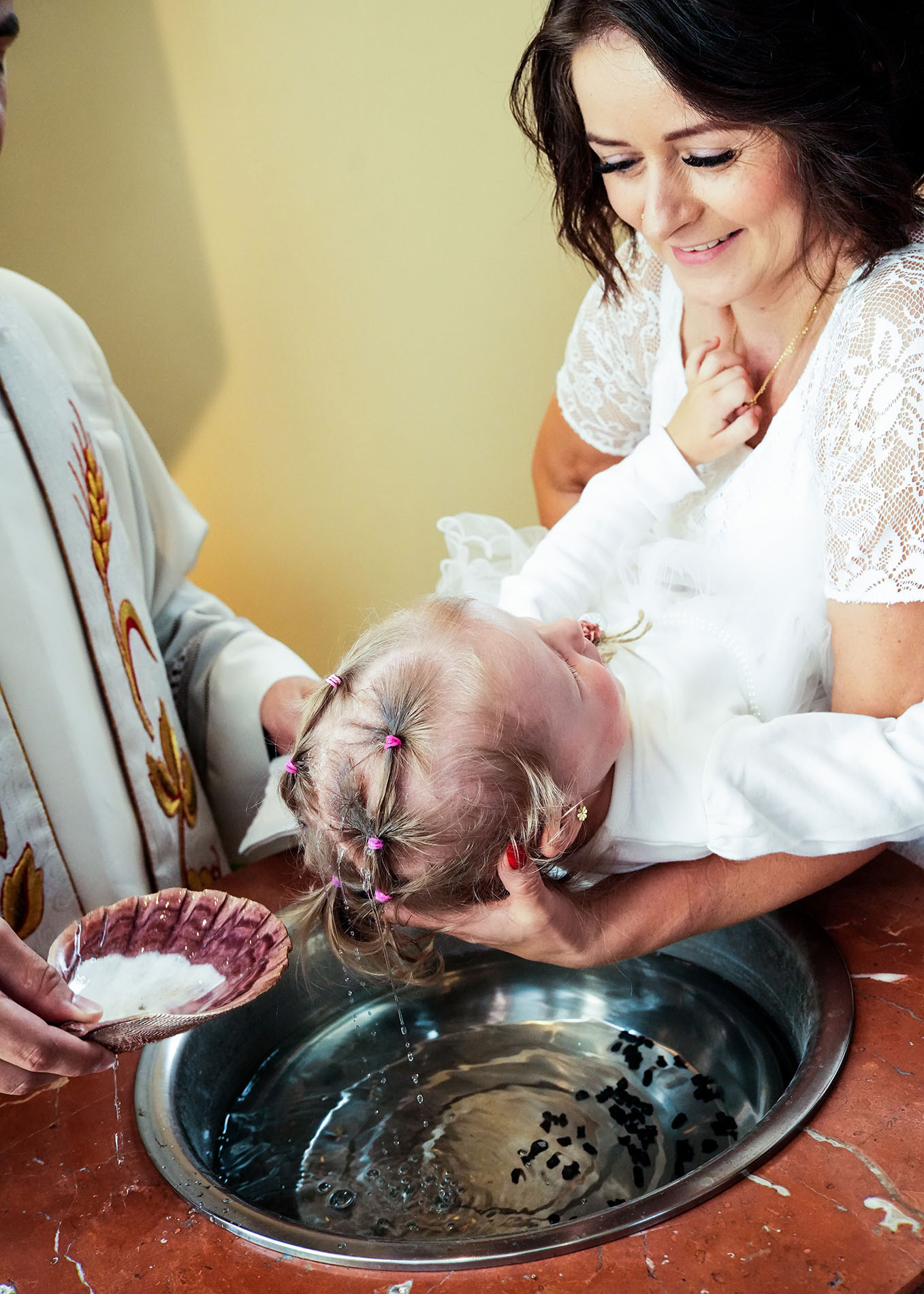 Ujęcie z góry przedstawia matkę trzymającą dziecko nad srebrną chrzcielnicą, podczas gdy ksiądz polewa wodą głowę dziecka z muszli. Matka uśmiecha się, a dziecko ma włosy spięte w małe kucyki...