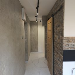 DANMONT Daniel Wilk - Korytarz po remoncie: gładzie na ścianach, marmurowe płytki podłogowe, ceglana ściana z przesuwnymi drzwiami i nowoczesne oświetlenie punktowe.