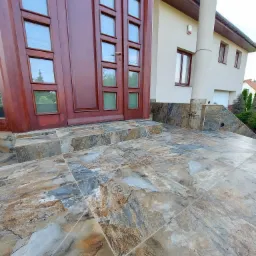 Wejście do domu z drewnianymi drzwiami z szybkami i schodami wyłożonymi płytkami imitującymi kamień w odcieniach szarości, brązu i beżu.