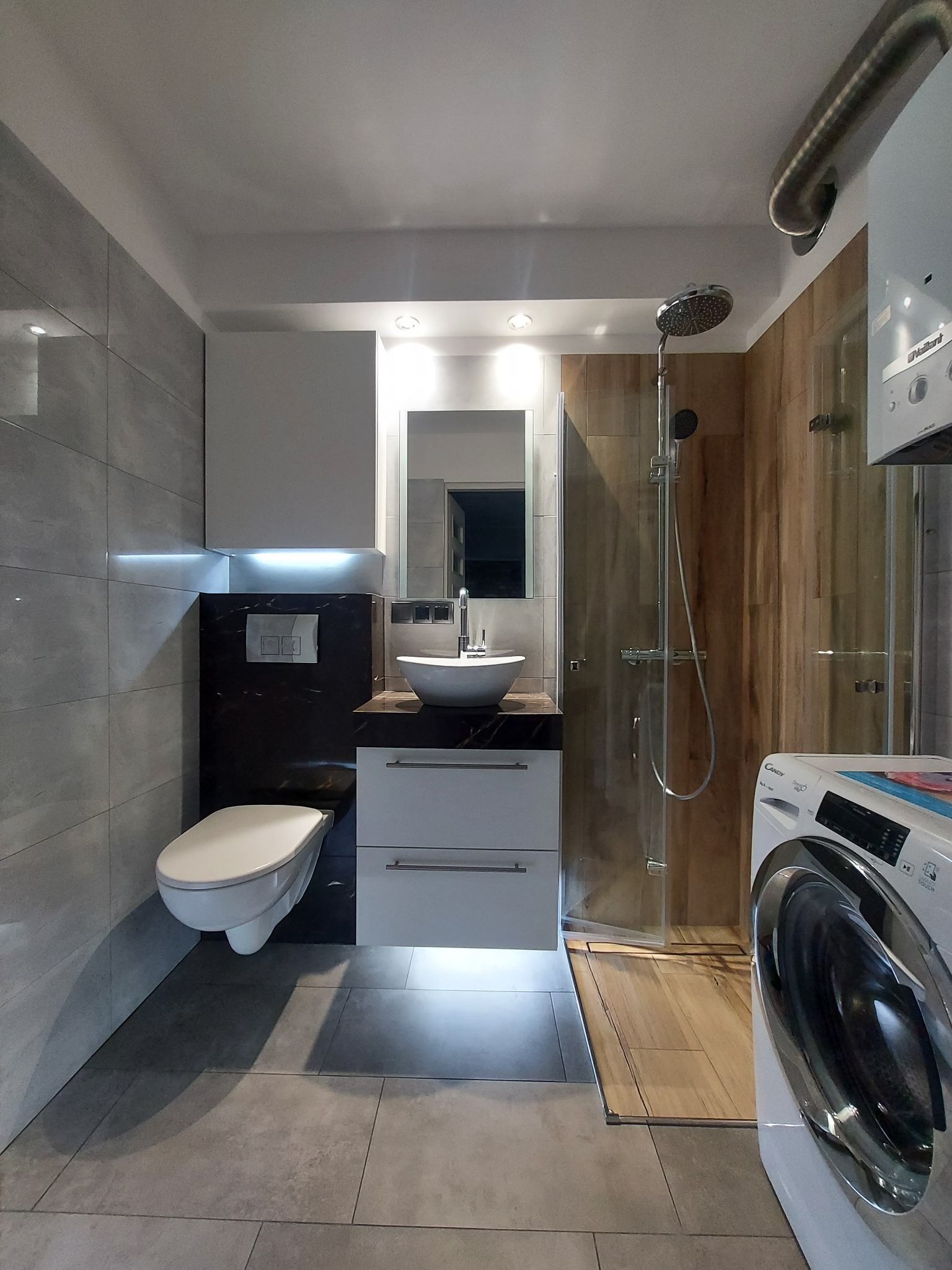 Nowoczesna łazienka z wiszącą toaletą, umywalką nablatową na szafce z szufladami, kabiną prysznicową z drewnopodobnymi płytkami oraz pralką Candy.