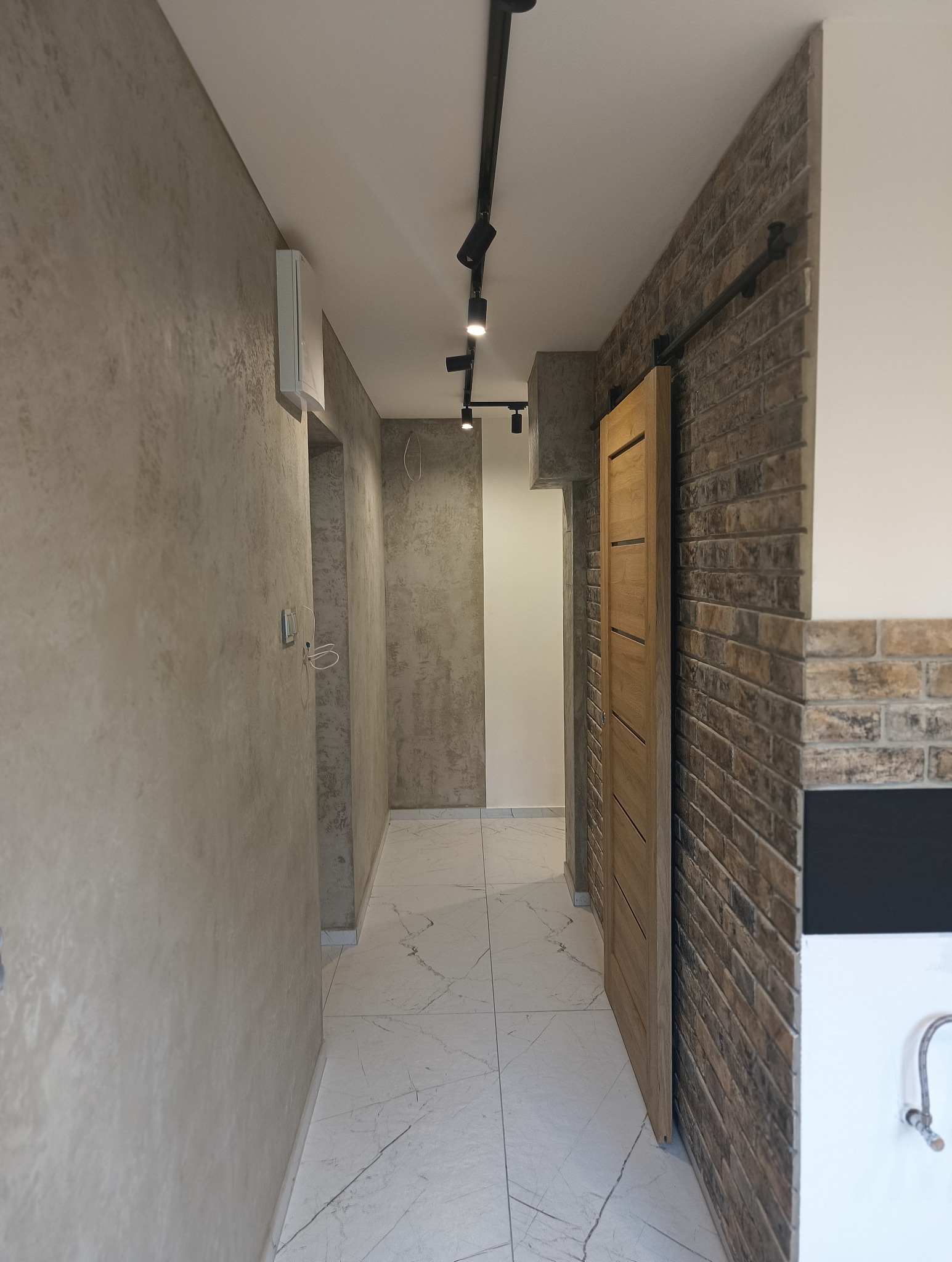 Korytarz po remoncie: gładzie na ścianach, marmurowe płytki podłogowe, ceglana ściana z przesuwnymi drzwiami i nowoczesne oświetlenie punktowe.