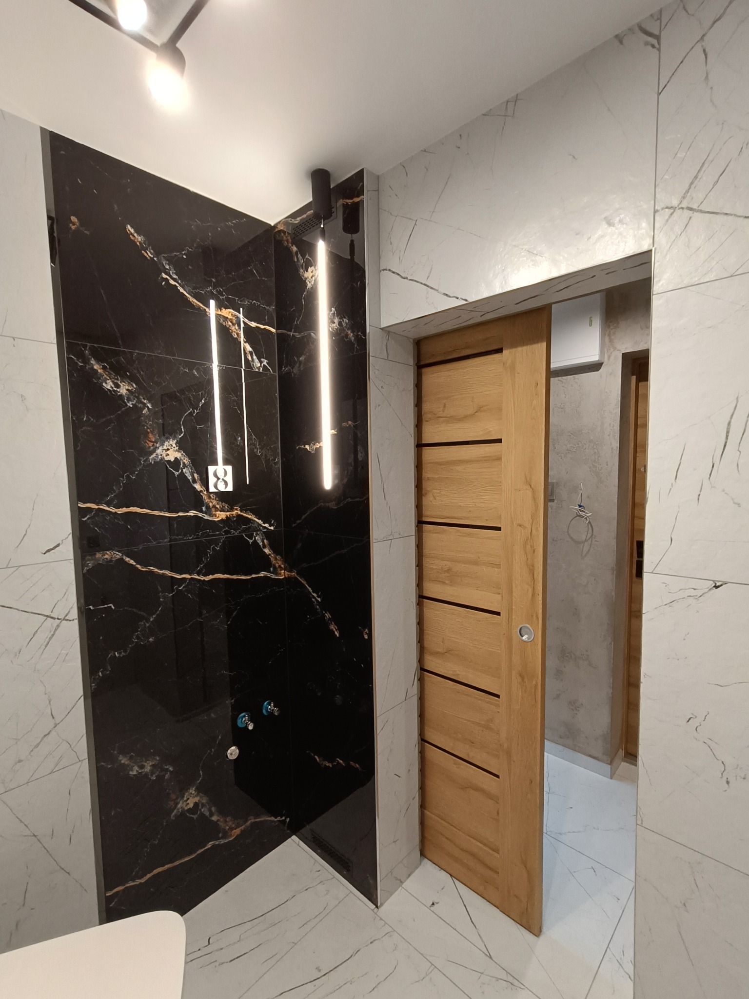 Nowoczesna łazienka z czarną ścianą prysznicową z marmurowym wzorem, oświetleniem LED i przesuwnymi drzwiami z jasnego drewna. Białe marmurowe płytki na ścianach i podłodze.