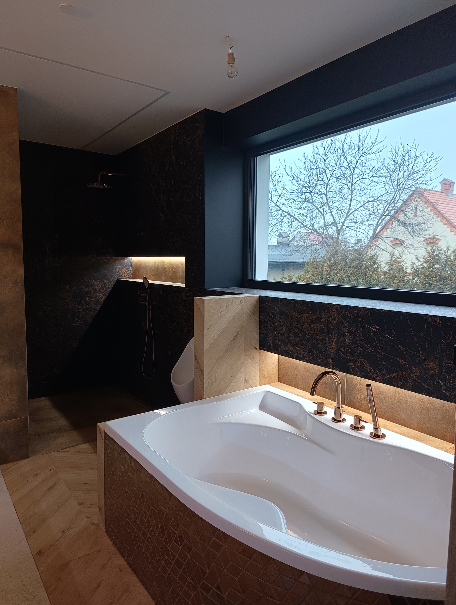 Nowoczesna łazienka z wanną, kabiną prysznicową i oknem. Ciemne płytki imitujące marmur, złota armatura, drewniane akcenty i podświetlenie LED tworzą luksusowy klimat.