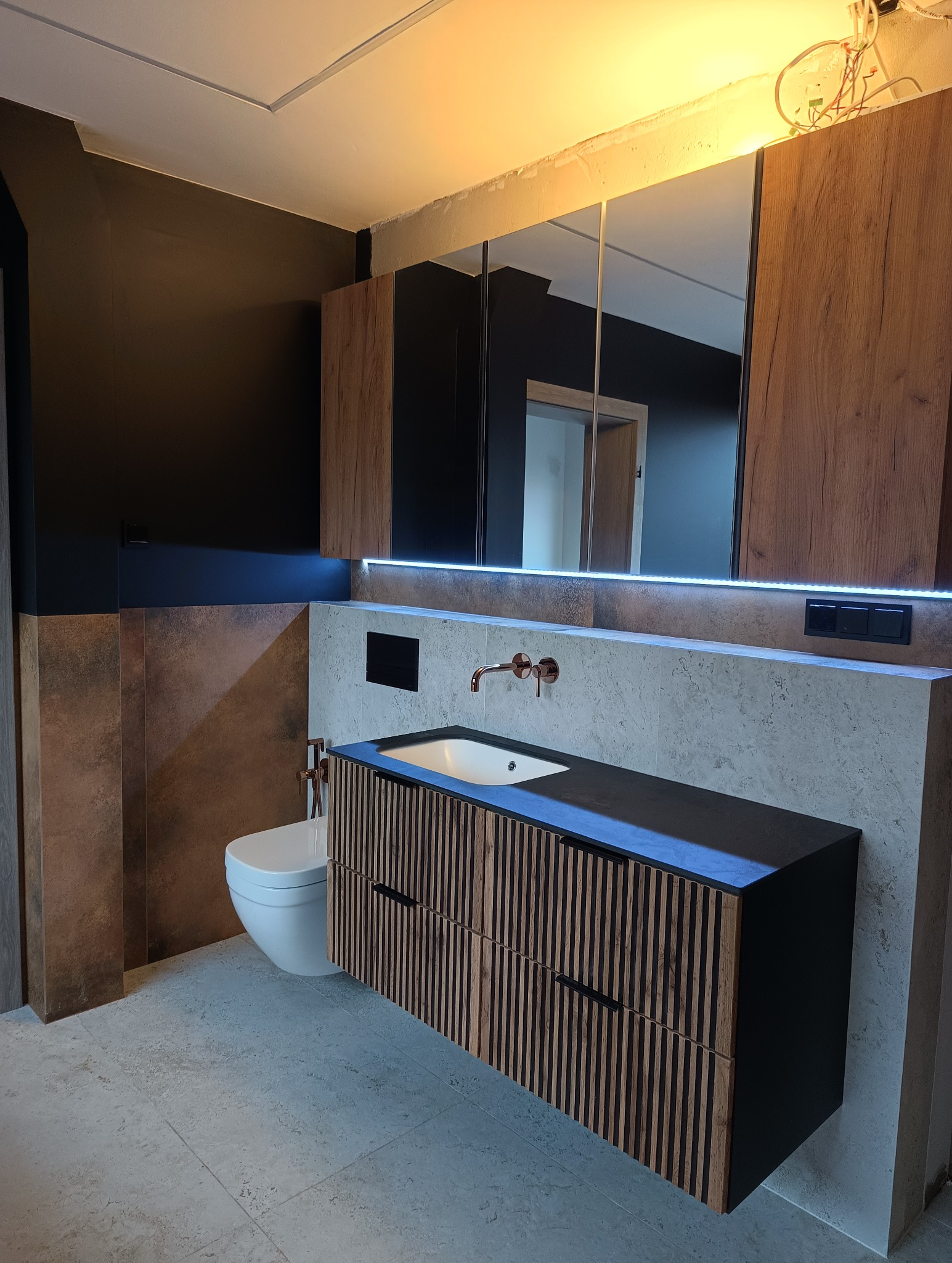 Nowoczesna łazienka po remoncie: czarne ściany, drewniane szafki z lamelami, podświetlane lustro i umywalka. Widoczne płytki imitujące kamień i wisząca toaleta.