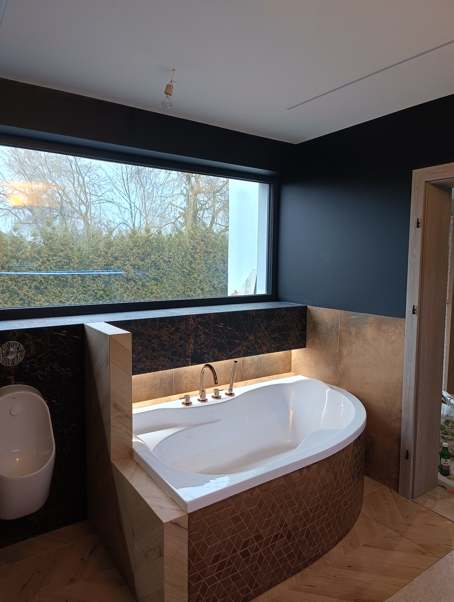 Nowoczesna łazienka z wolnostojącą wanną obudowaną mozaiką, marmurową umywalką i dużym oknem z widokiem na zieleń. Minimalistyczny design, eleganckie wykończenie.