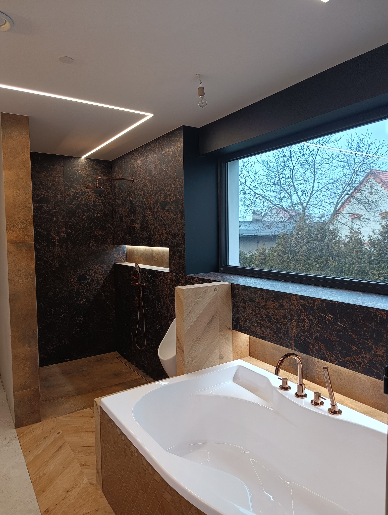 Nowoczesna łazienka z kabiną prysznicową typu walk-in, białą wanną i armaturą w kolorze miedzi. Ciemne płytki imitujące marmur i oświetlenie LED tworzą elegancki klimat.