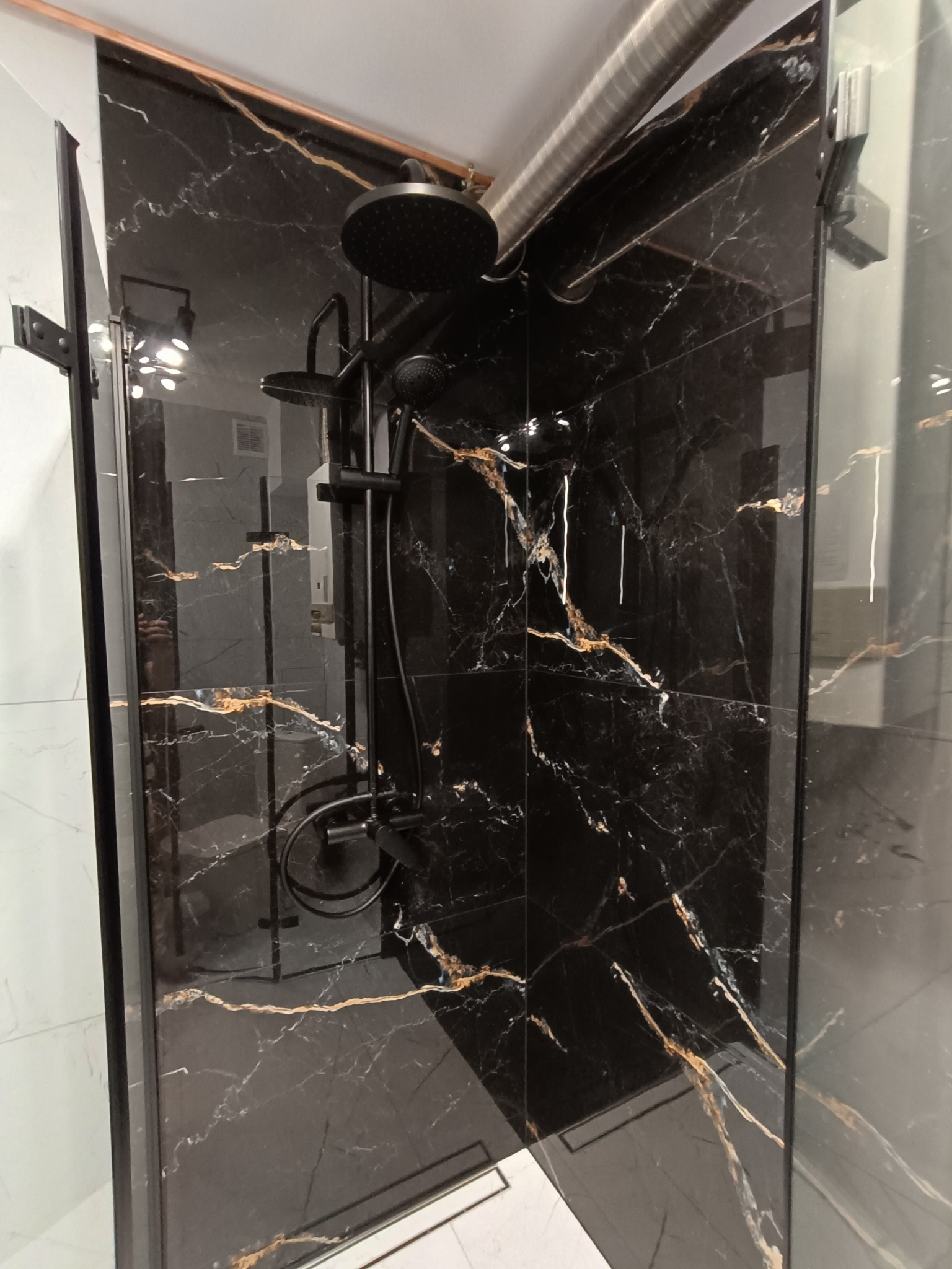 Kabina prysznicowa z czarnymi płytkami imitującymi marmur z złotymi żyłami i czarną armaturą. Widoczny fragment podłogi z białych płytek.