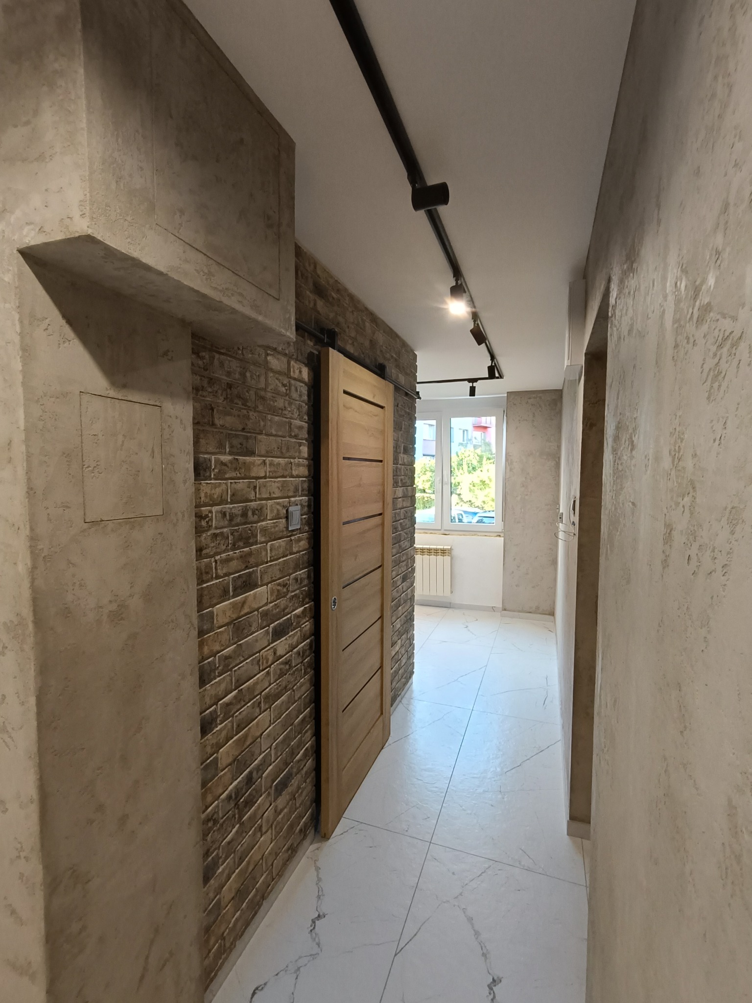 Korytarz w stylu loft: cegła dekoracyjna, betonowe tynki, drewniane drzwi przesuwne, oświetlenie szynowe i marmuropodobna posadzka. Widok z perspektywy.