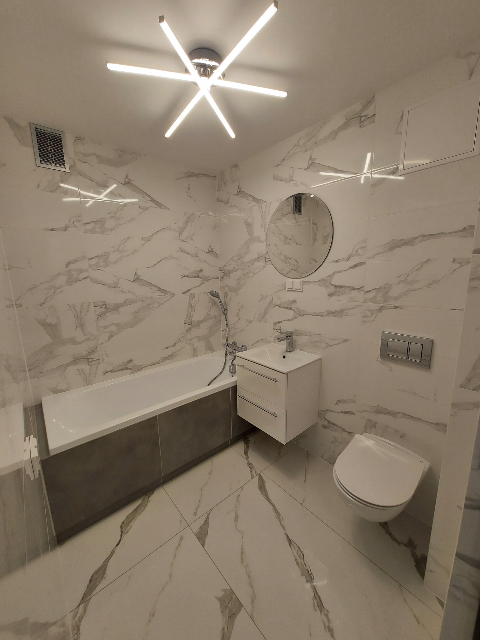 Nowoczesna łazienka z marmurowymi płytkami, podwieszaną toaletą, umywalką z szafką i okrągłym lustrem, oświetlona designerską lampą sufitową.