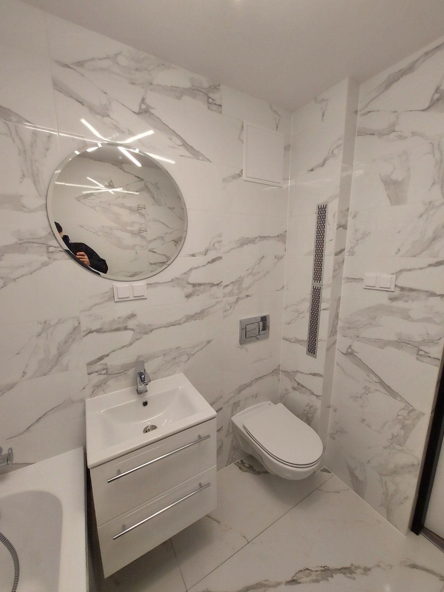 Nowoczesna łazienka wykończona białymi płytkami imitującymi marmur, z okrągłym lustrem z podświetleniem LED, wiszącą toaletą i szafką pod umywalkę z dwiema szufladami.