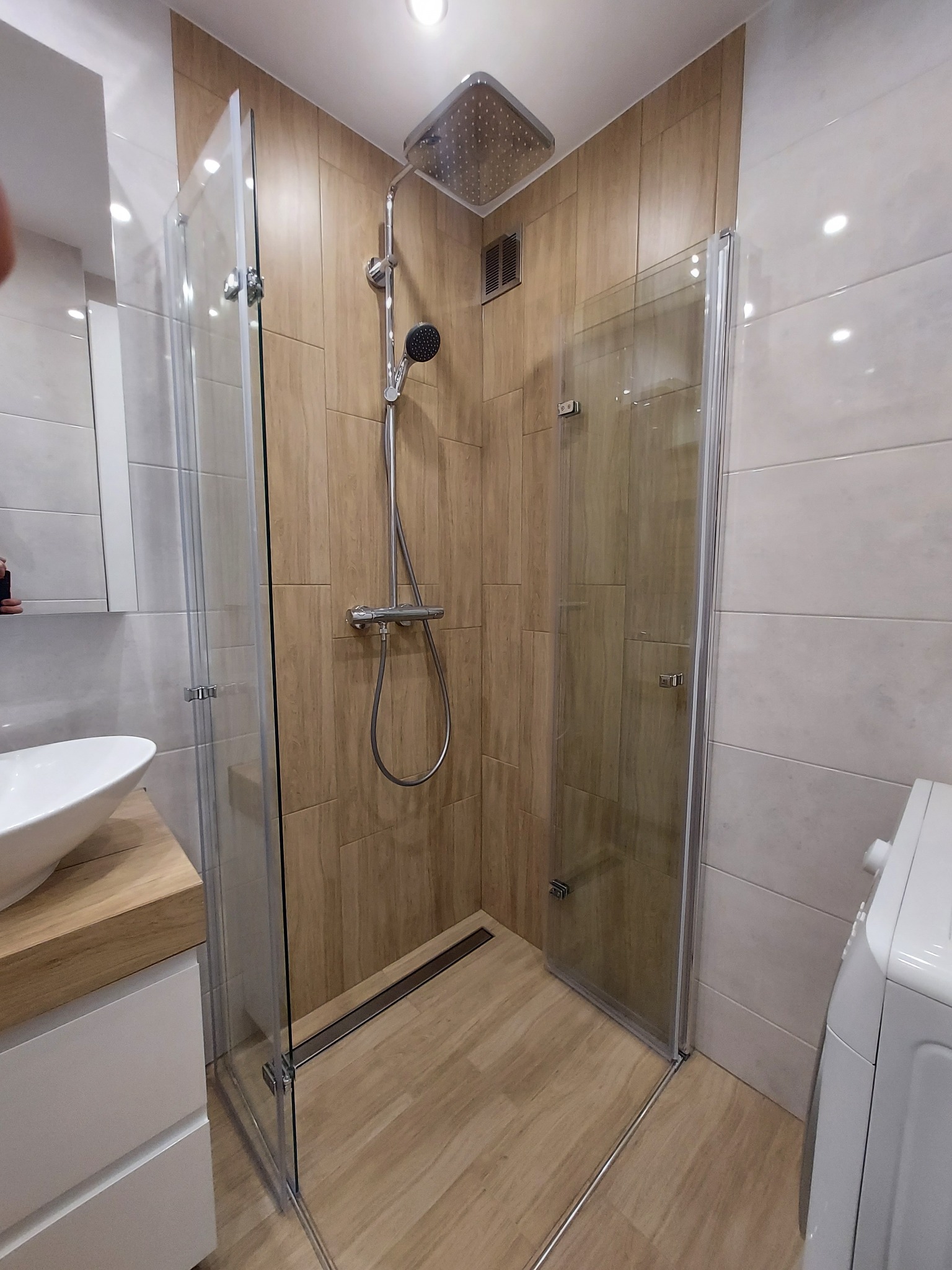 Nowoczesna łazienka z kabiną prysznicową typu walk-in, wykończona płytkami imitującymi drewno, z odpływem liniowym w podłodze i armaturą w kolorze chromu.