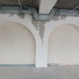 Wykończmy To - Dwa łukowate wnęki w ścianie, w trakcie prac wykończeniowych, widoczne ślady po gładzi szpachlowej, sufit z instalacją wentylacyjną, podłoga zabezpieczona folią.