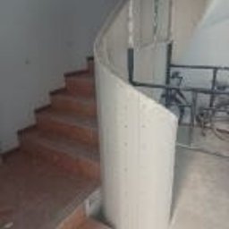 Wykończmy To - Klatka schodowa w trakcie budowy, widoczne schody z drewnianymi stopniami, obok kręta ściana z płyty kartonowo-gipsowej na metalowym stelażu, surowy stan wykończenia.